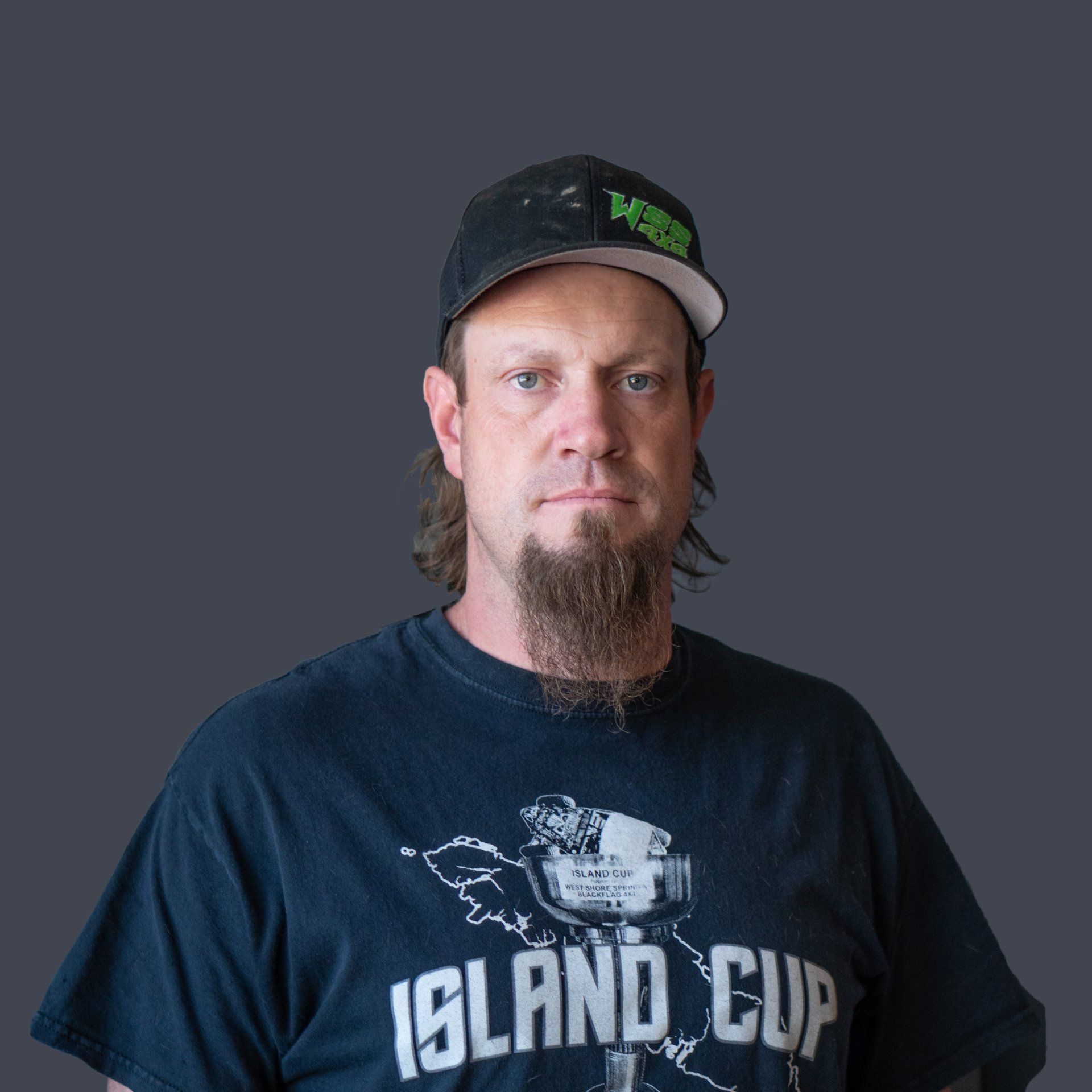 Man with goatee wearing a baseball cap and t-shirt, standing against a gray backdrop.