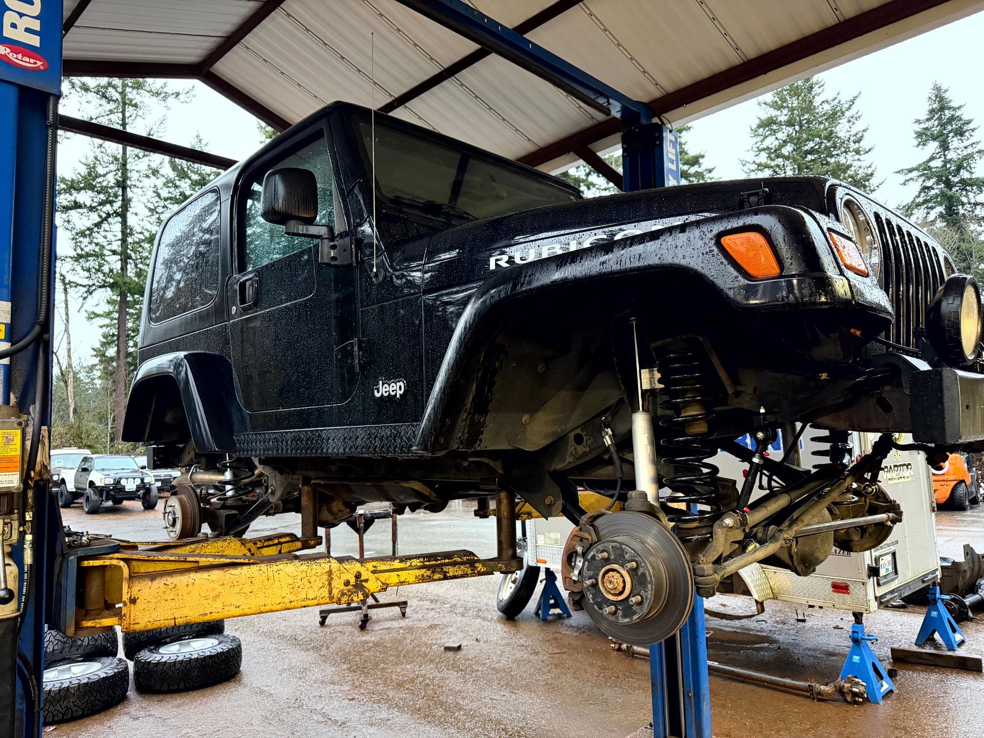 Black Jeep Wrangler on a lift in a repair shop, undergoing maintenance; wheels removed.