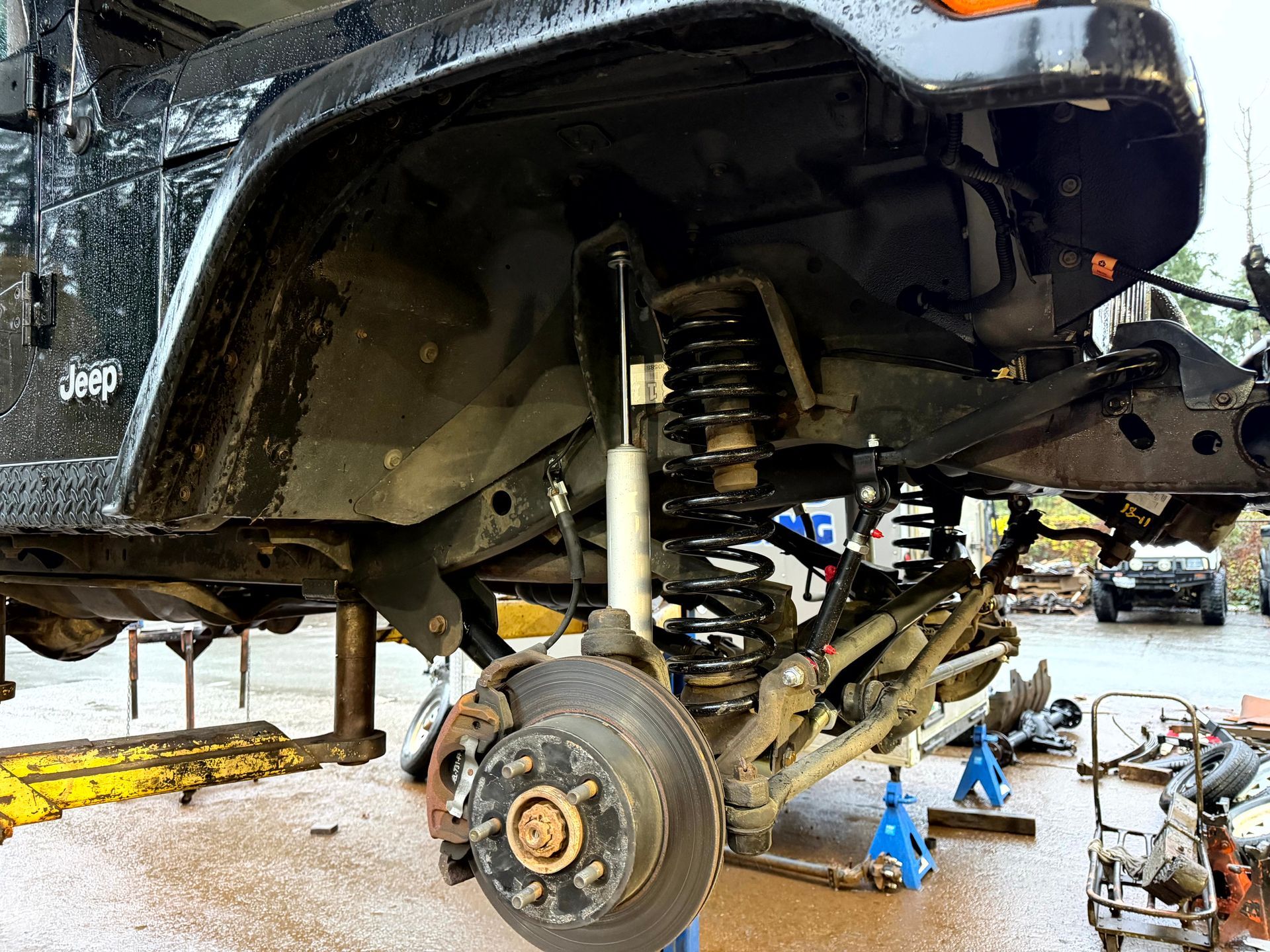 Black Jeep's front suspension disassembled; tire removed. Supported by jack stands. Mechanic working outdoors.