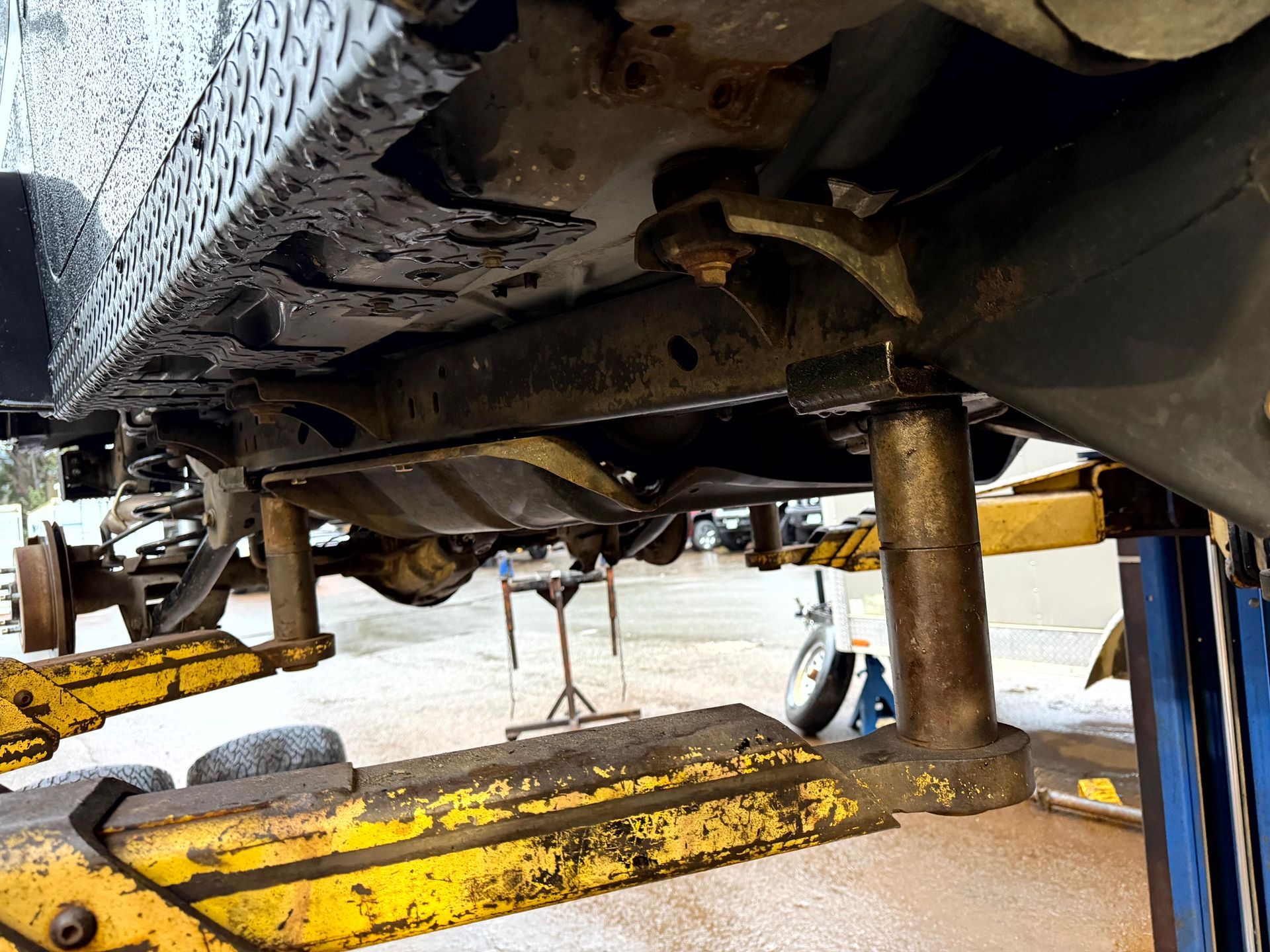 Underside of a vehicle lifted on a yellow hoist; shows undercarriage with rust and mechanical components.