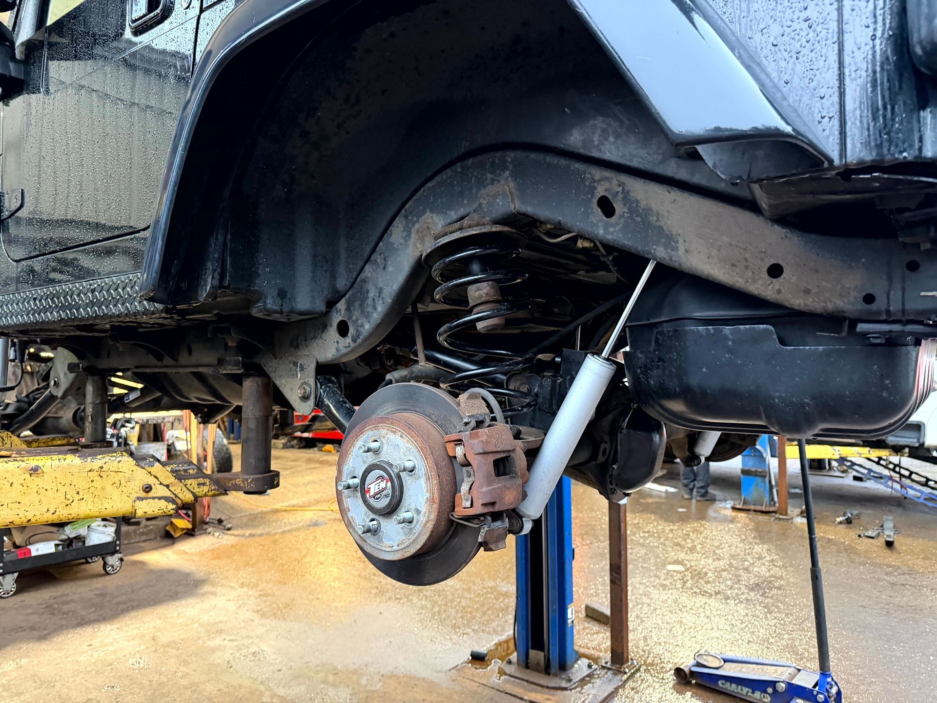 Black Jeep rear wheel raised on a lift for repair. Rusty brake rotor and shock absorber visible.