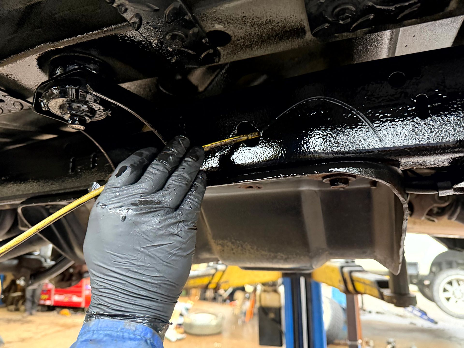 Gloved hand using a wand to apply black coating to the underside of a vehicle in a garage.