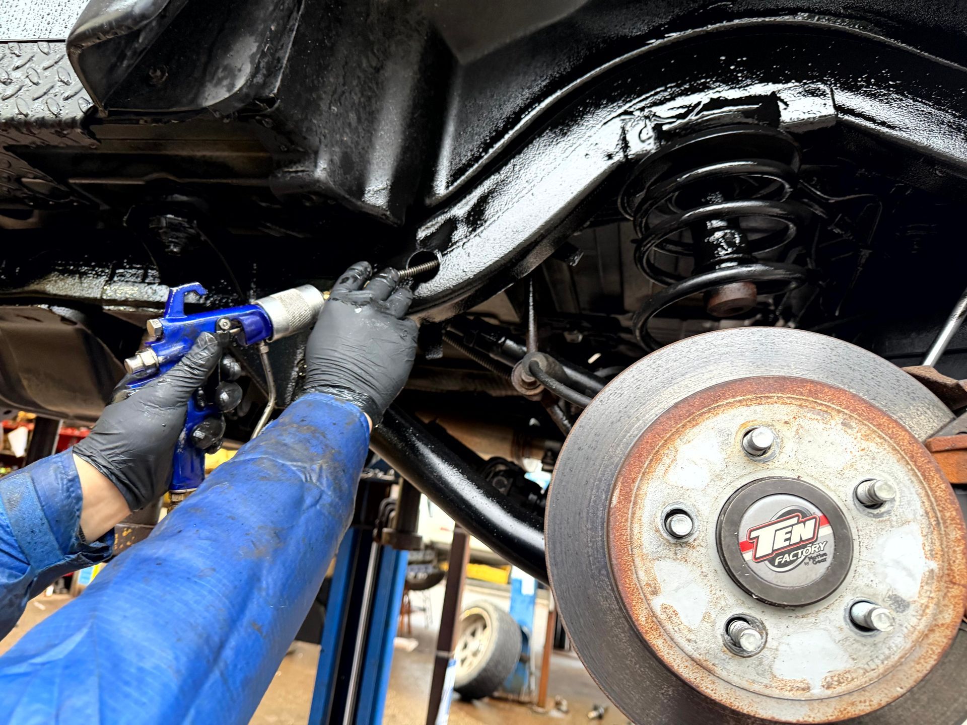 Person in blue coveralls sprays black coating on vehicle undercarriage near a disc brake.