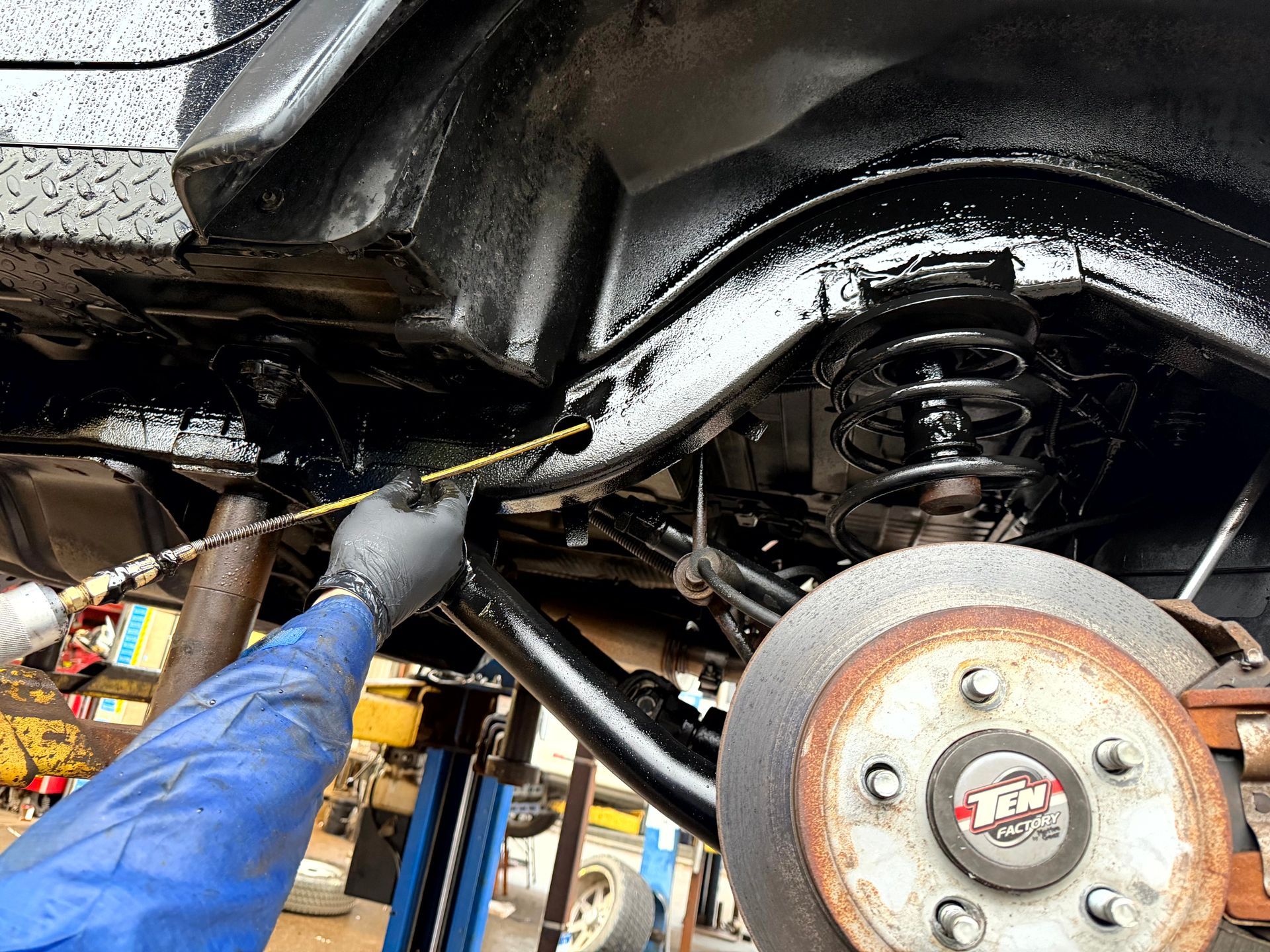 Person spraying black undercoating on a vehicle's undercarriage in a repair shop.