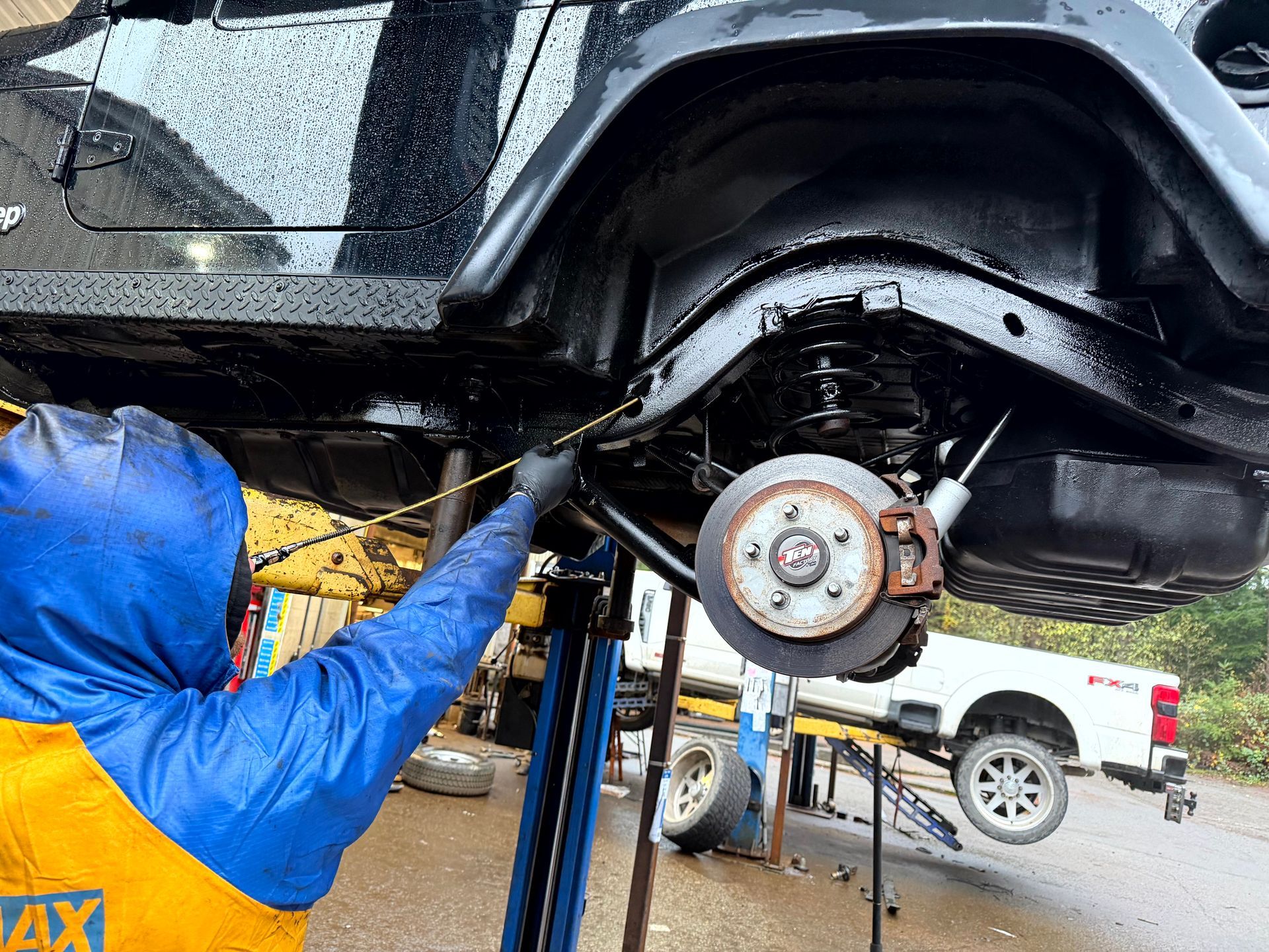 Person in blue rain gear working on a black Jeep lifted on a hoist.