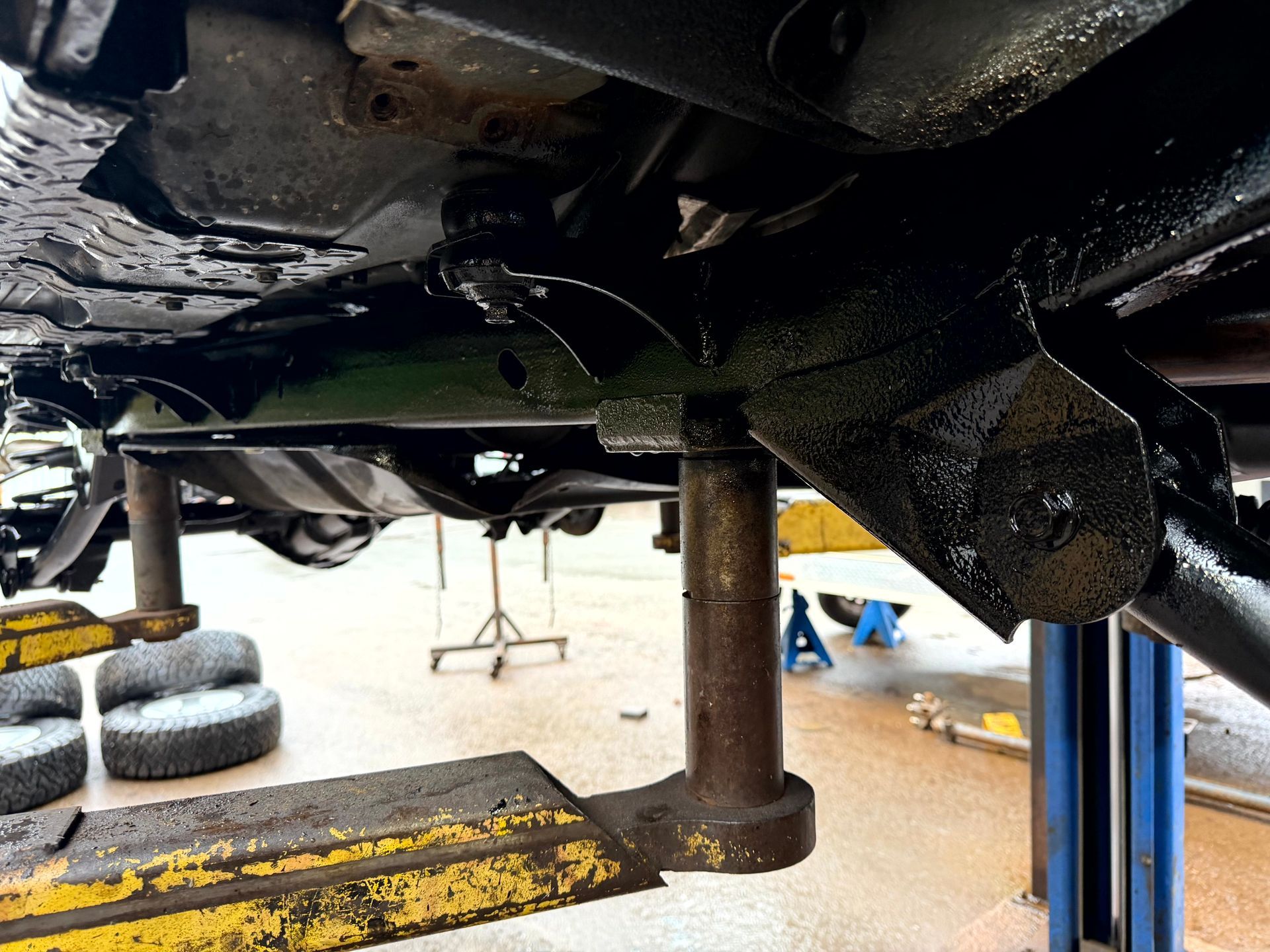 Undercarriage of a vehicle raised on a jack stand, showing the frame, suspension components, and the jack.