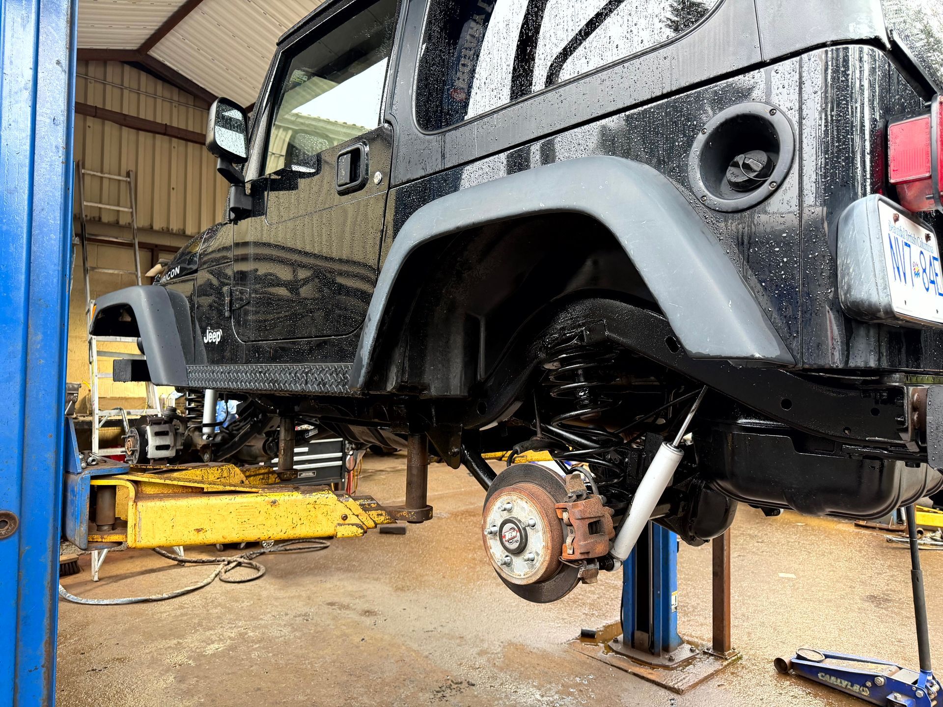 Black Jeep Wrangler on a lift in a garage, rear wheel off, showing suspension components.