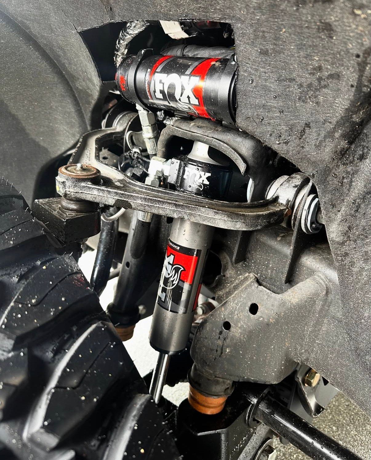 Close-up of a truck suspension showing Fox shocks and coil-overs, black and red against a gray wheel well.