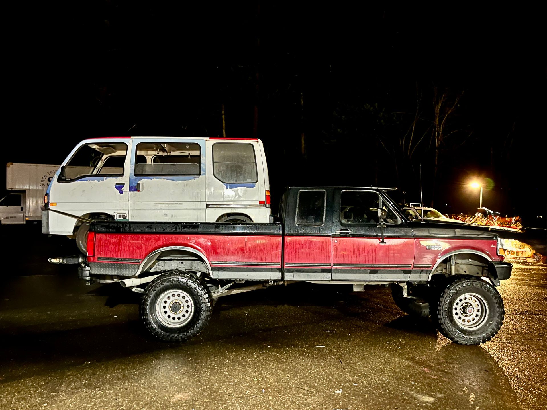 Red lifted pickup truck towing a small white van in a dark, outdoor setting.