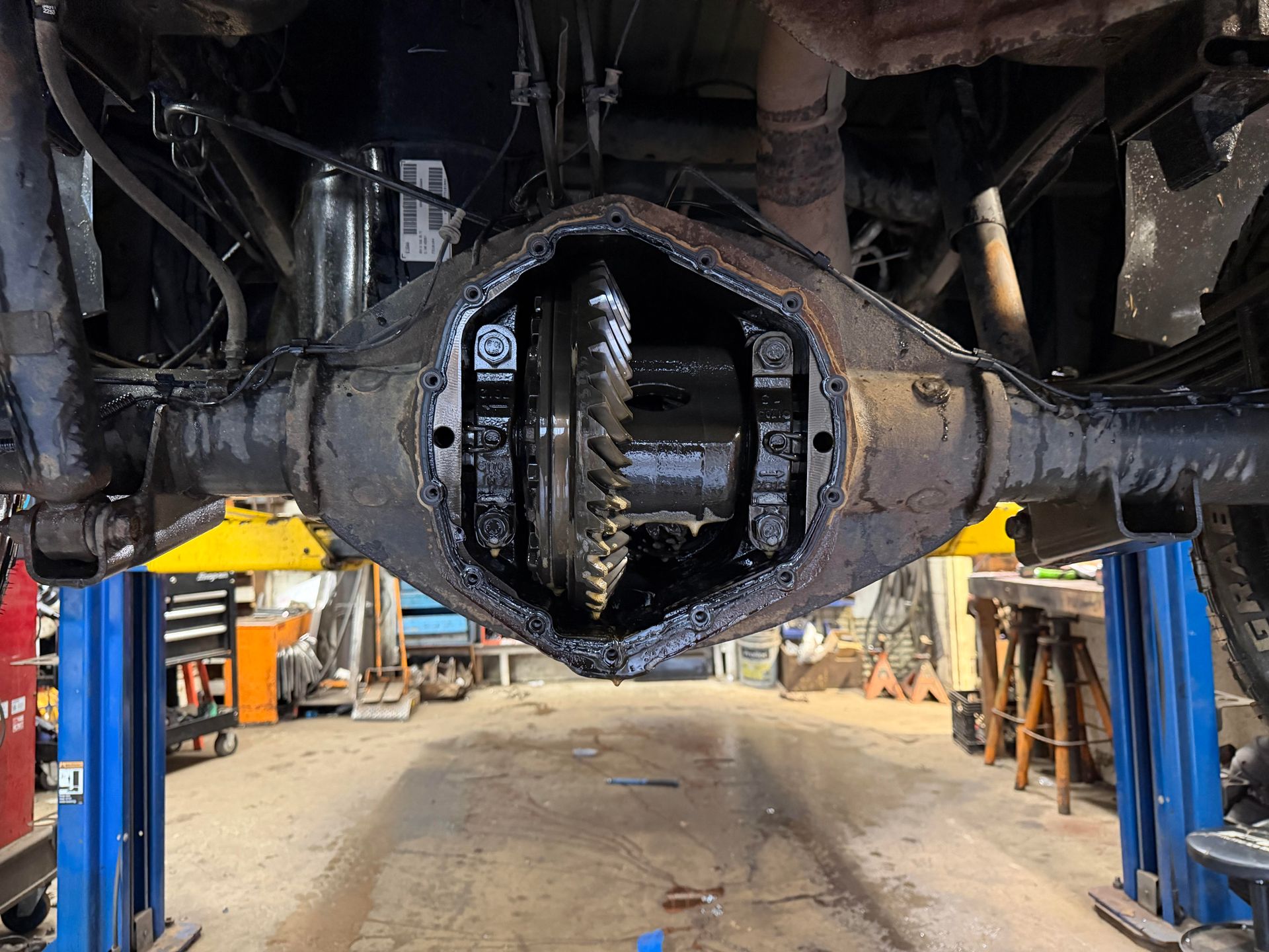 Open rear differential on a vehicle, showing internal gears, covered in oil. Workshop setting.