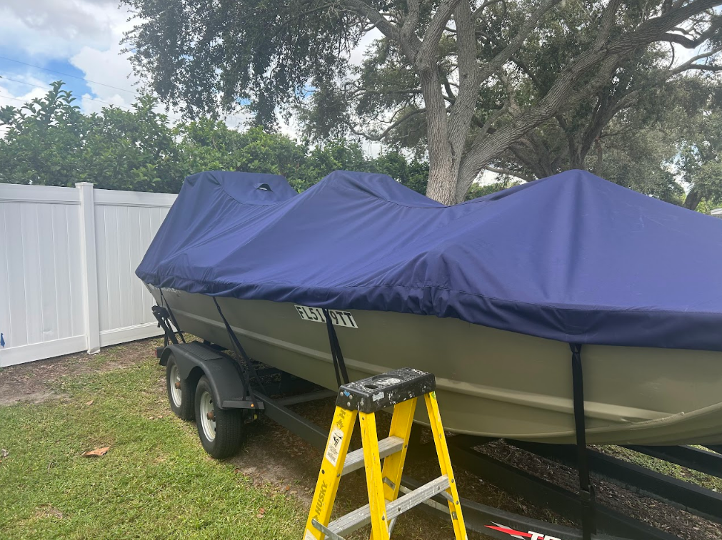 Boat on a trailer, covered with a blue tarp, beside a white fence and a tree, with a yellow ladder.