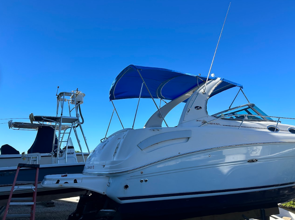 Blue-canopied motorboat on land, sunny day. Boat is white with blue trim. Another boat and tower in background.