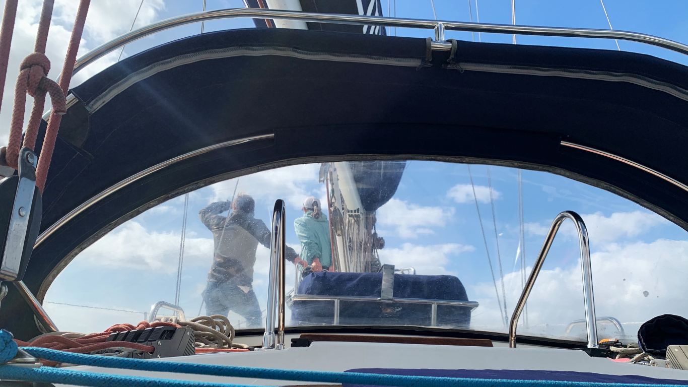People on sailboat deck, under a black canvas canopy, working near the mast; blue sky.