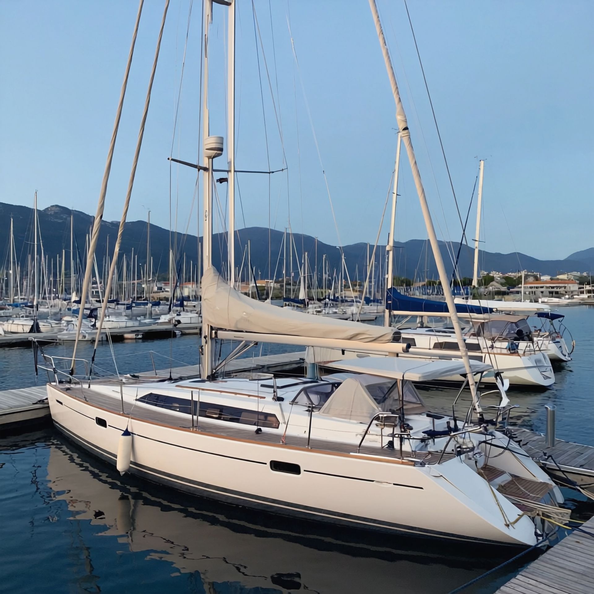 Sailboat docked in a marina, mountains in the background. The boat is white, the setting is sunny.