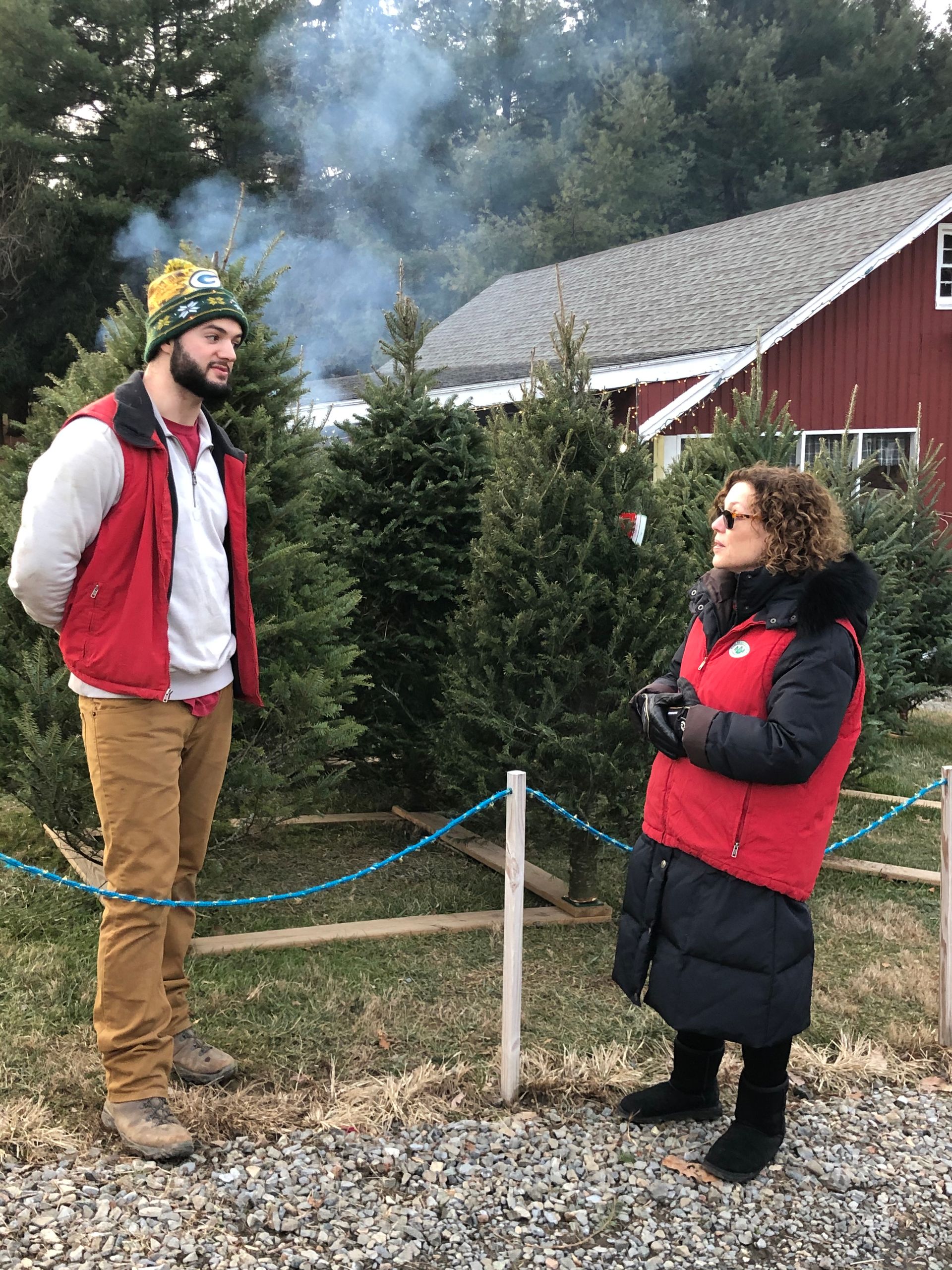 A man and a woman are standing next to each other in front of a christmas tree farm.