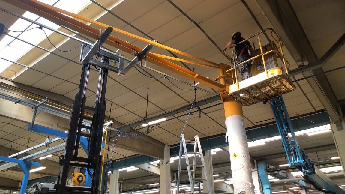 Magazzino industriale con un operaio su una piattaforma elevatrice gialla intento a riparare macchinari e travi in ​​quota.