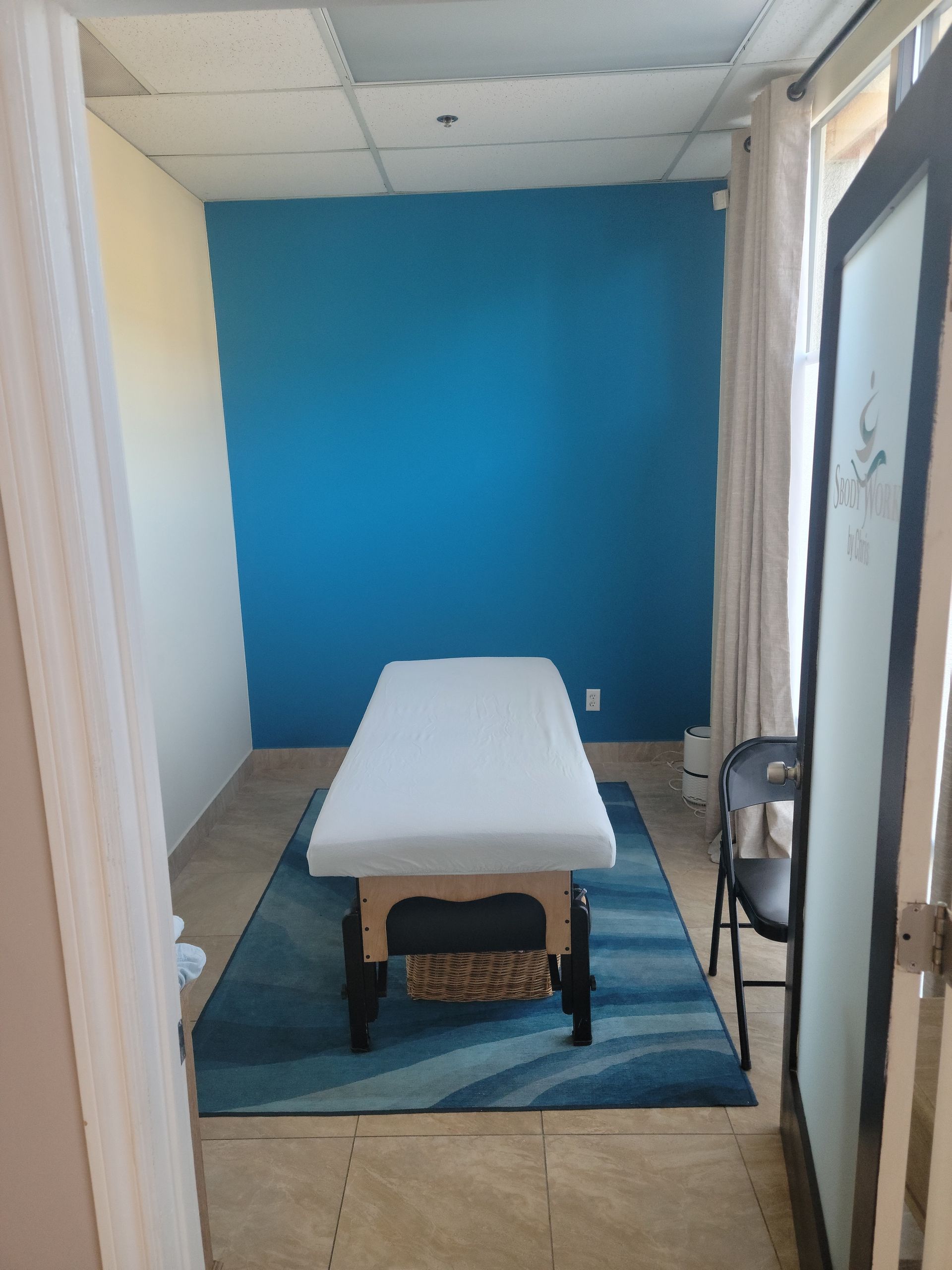 Small treatment room with a white massage table, blue back wall, and striped rug.