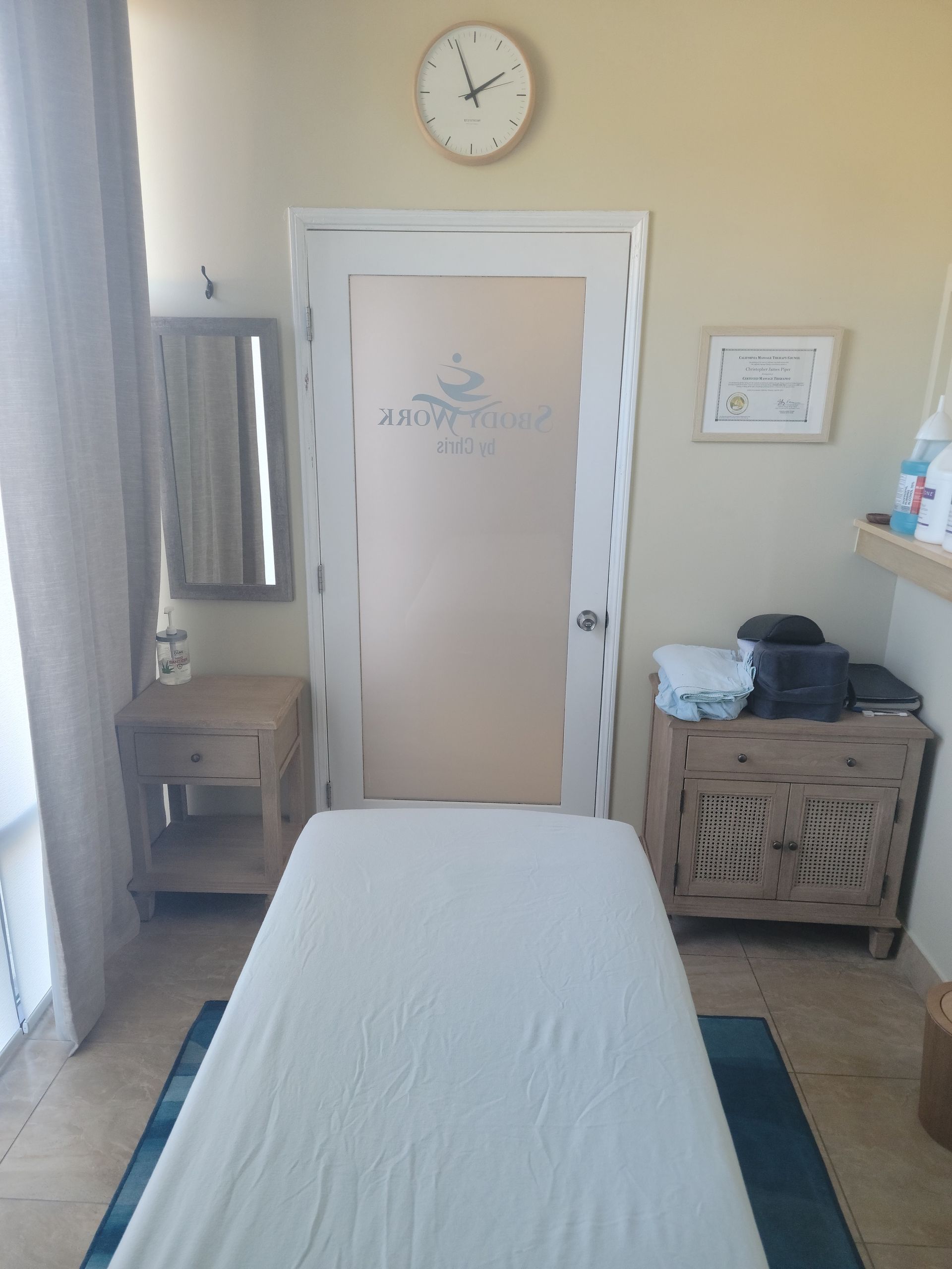 Massage room with treatment table, wall clock, mirror, cabinet, and towels by the door