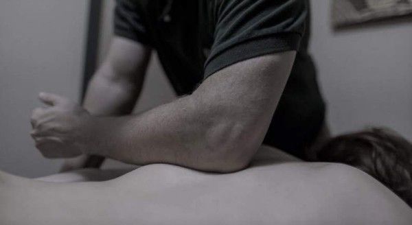 Person in a black shirt leaning over another person lying face down on a surface, in a dim room.