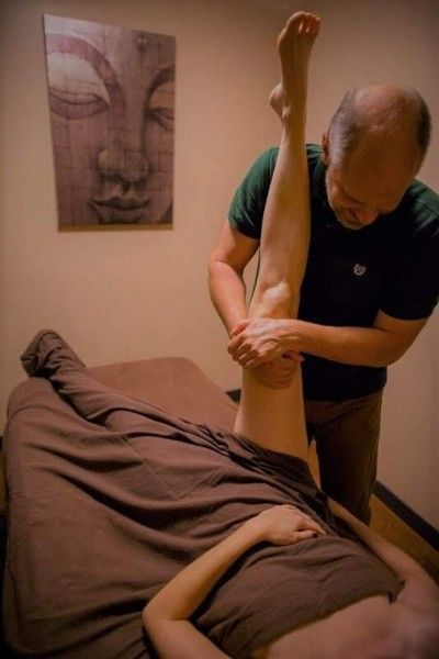 Massage therapist stretching a client’s leg in a treatment room, with a wall art portrait nearby.