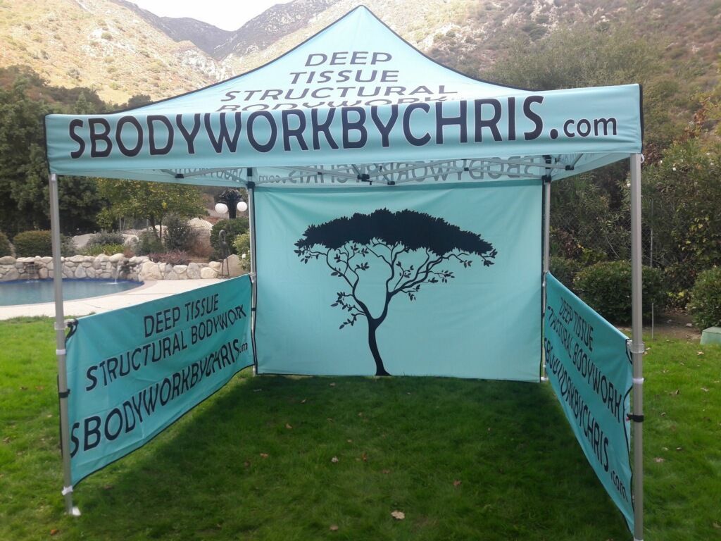 Blue tent booth with “SBodyworkbyChris.com” signs and a tree graphic on a grassy outdoor path.