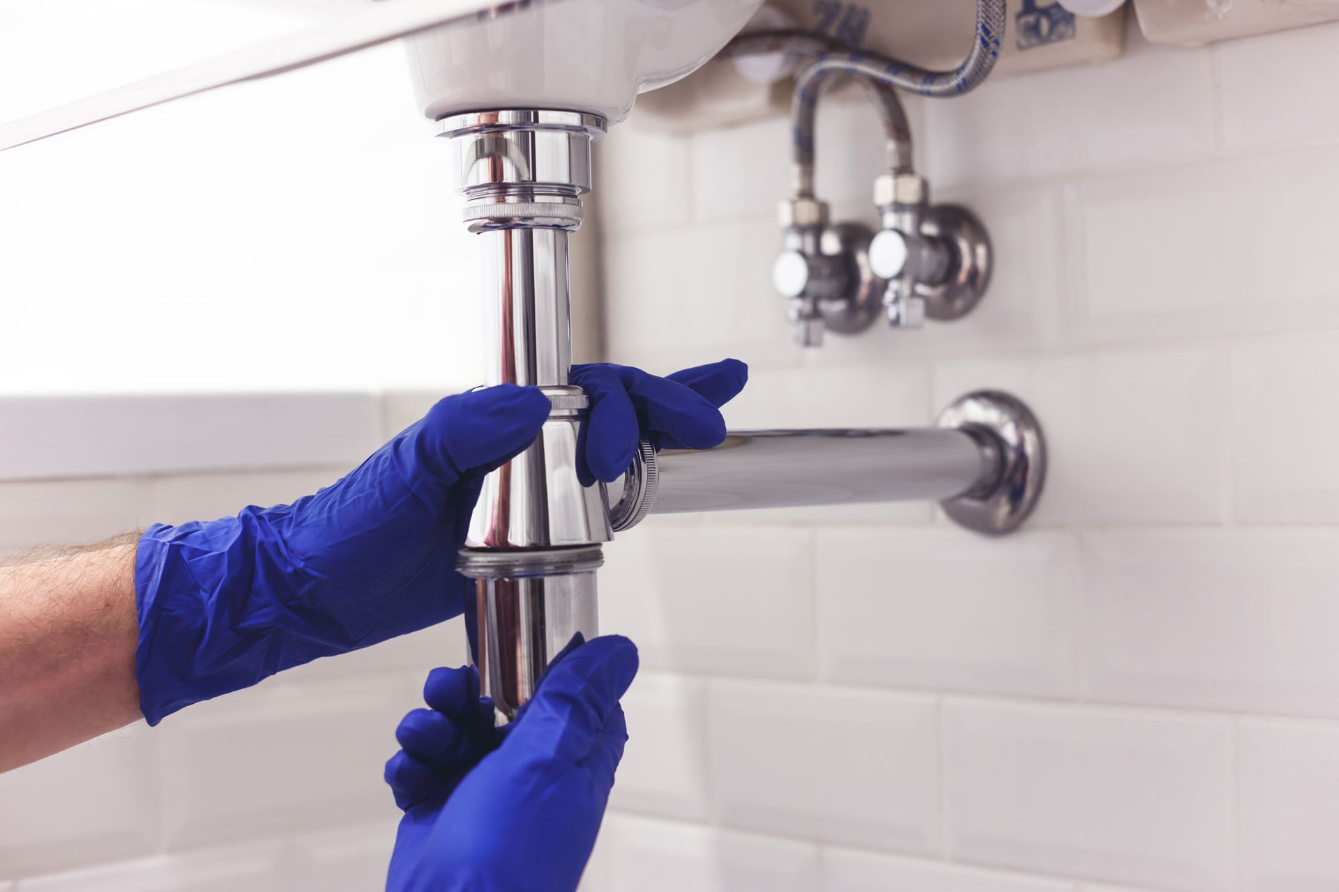 Plumber’s gloved hands tightening a sink drain pipe under a bathroom sink. Plumber’s gloved hands tightening a sink drain pipe under a bathroom sink.