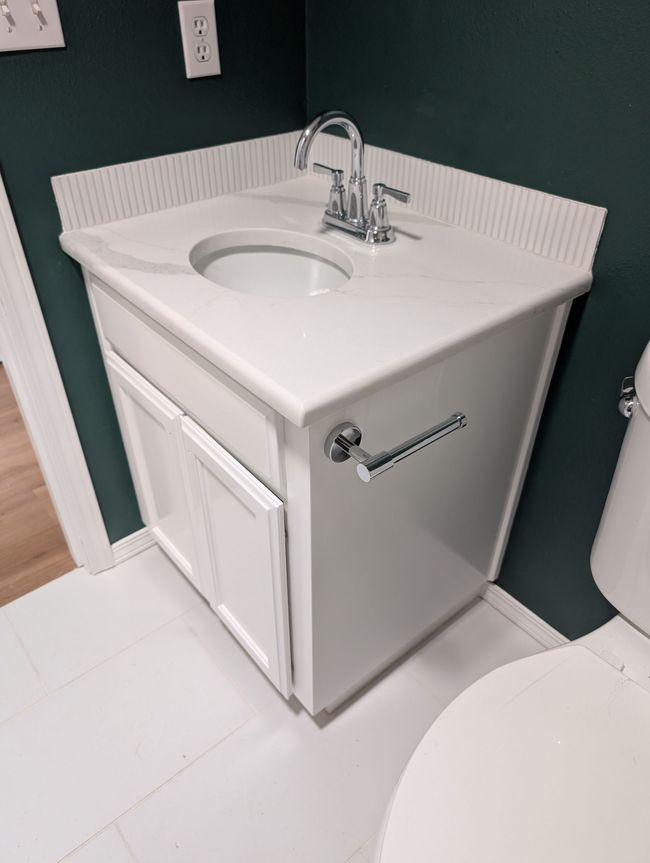 White corner bathroom sink vanity with chrome faucet and towel bar against green walls and white tile floor