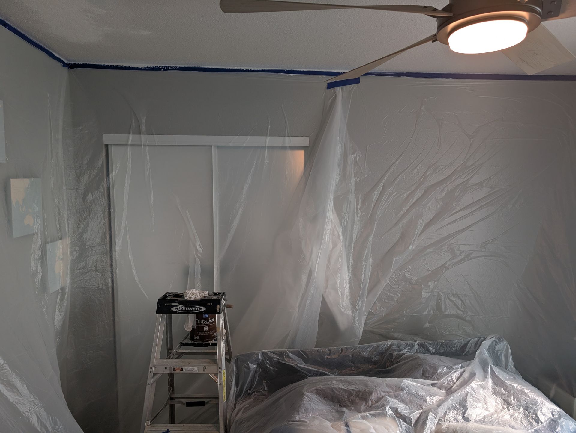 Room covered in plastic sheeting, with a lit ceiling fixture, a fan, and covered furniture during renovation.