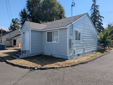 Small light-gray house with white trim and a side yard beside a paved road on a sunny day