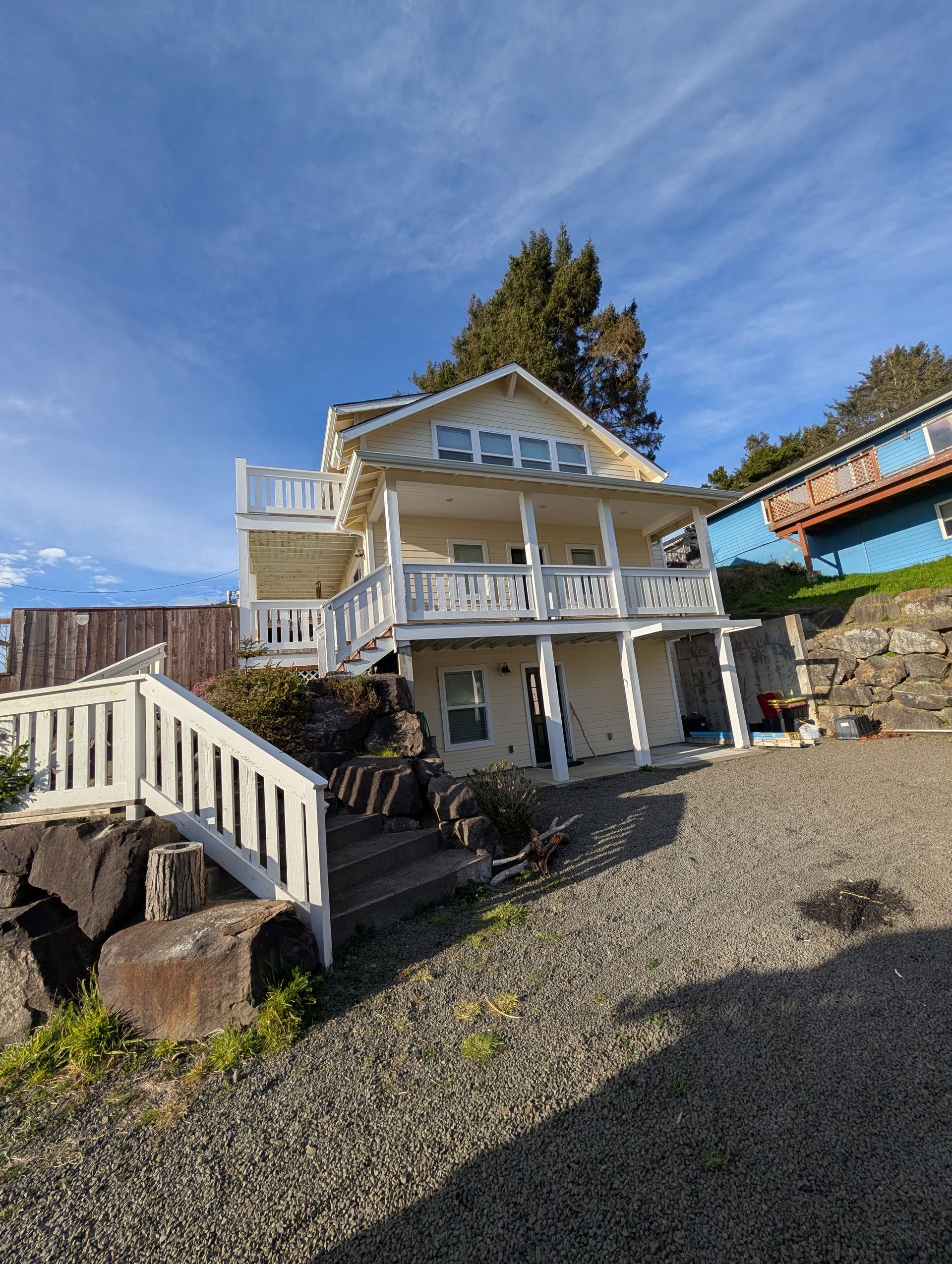White seaside house with wraparound porches on a gravel slope under a blue sky