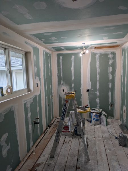 Narrow room under renovation with drywall patches, unfinished plank floor, ladder, tools, and a window on the left