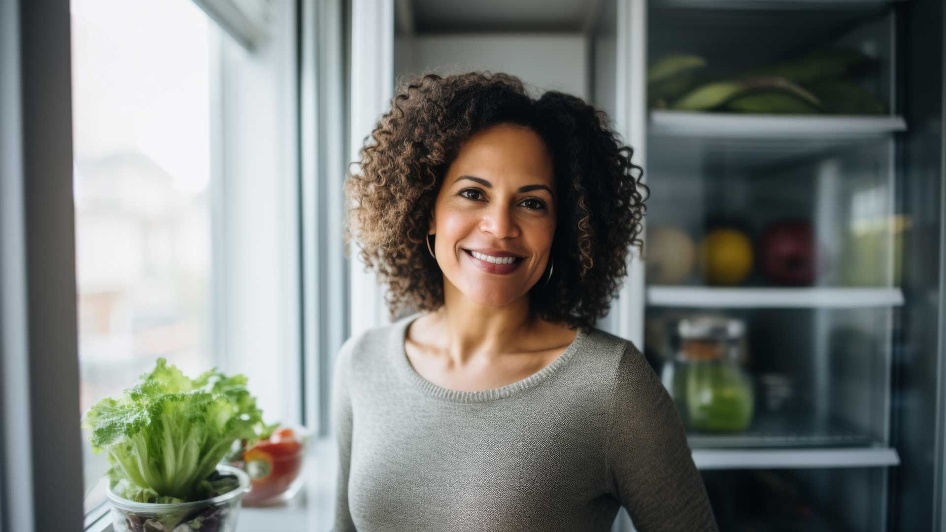 Smiling woman with curly hair stands near window with lettuce and open refrigerator with produce.