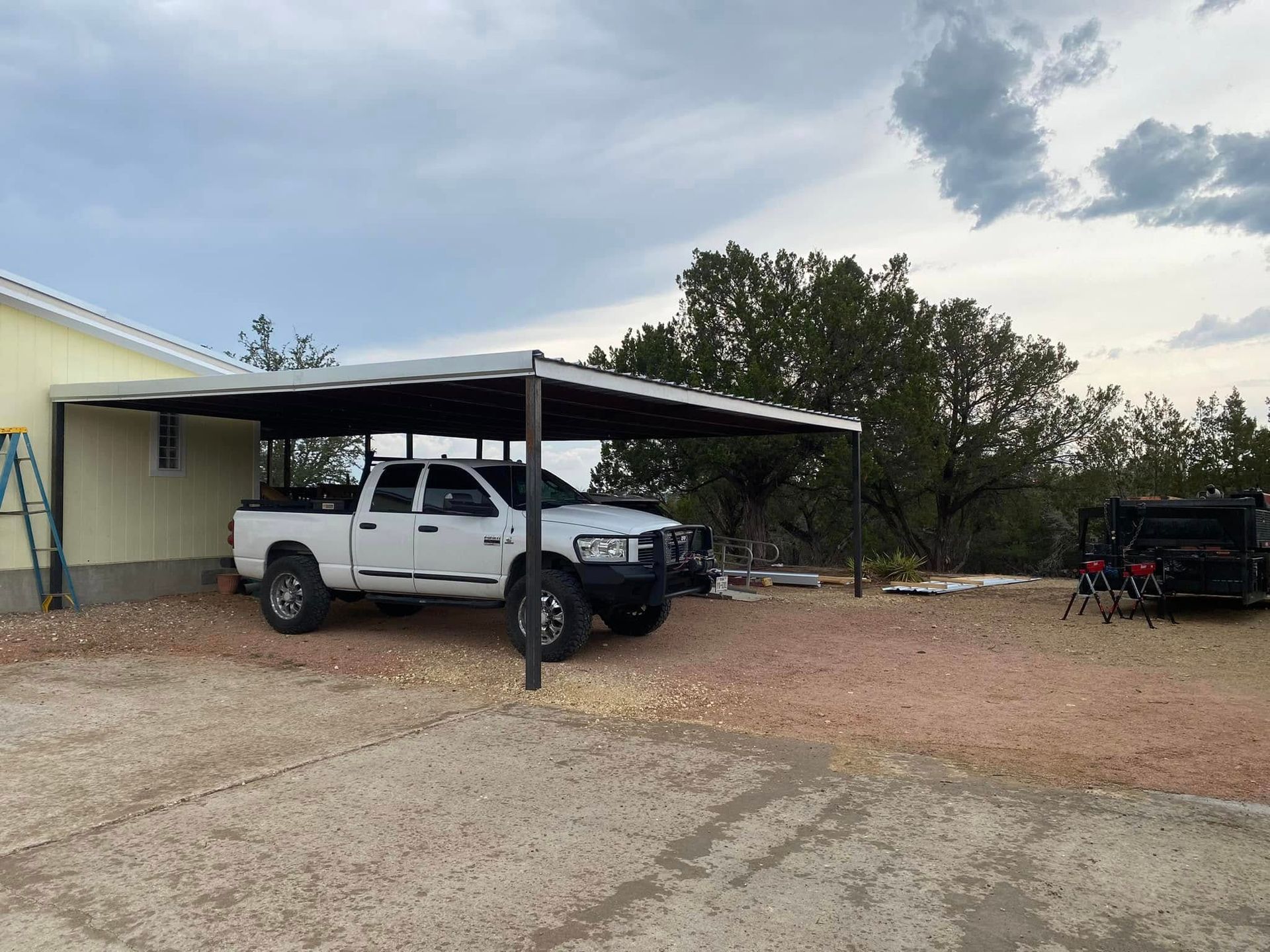 White pickup truck under a carport beside a beige building on a gravel driveway with trees and a trailer nearby