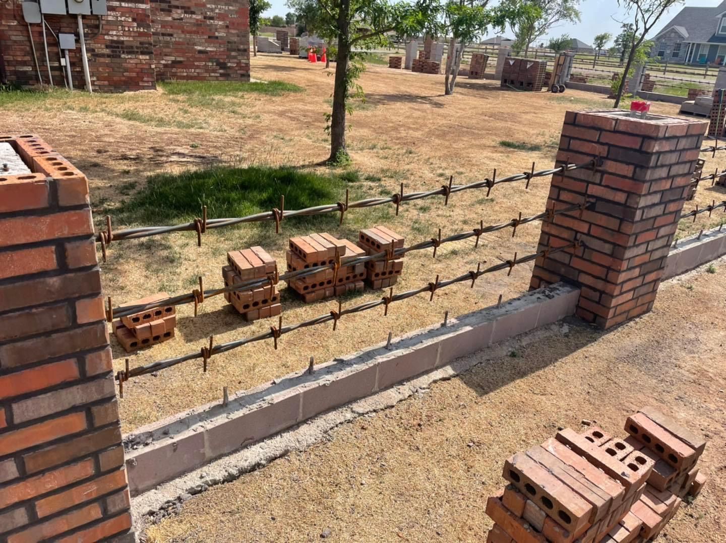 Brick wall under construction with bricks, wire fencing, and unfinished pillars in a dry yard