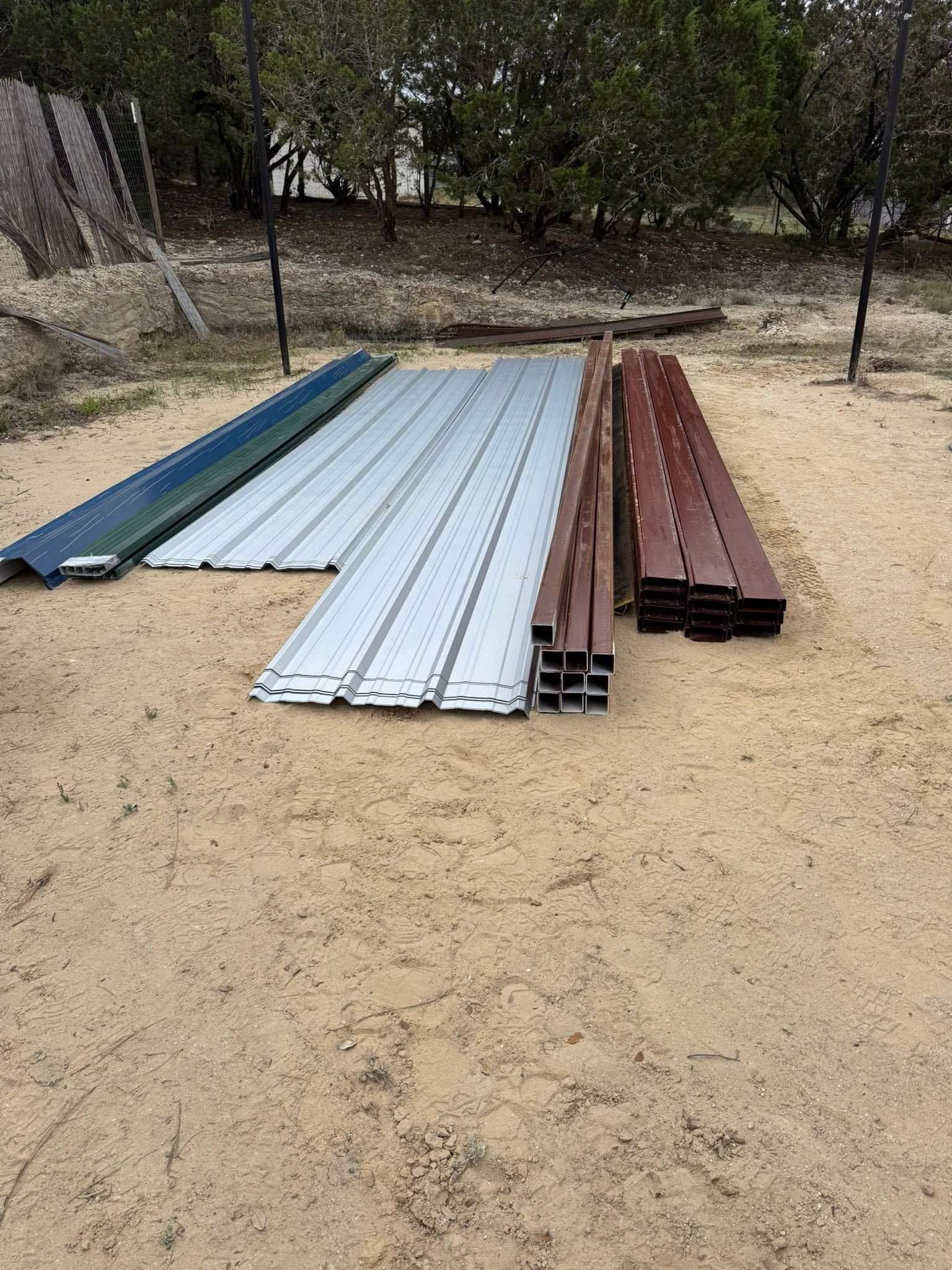 Stacks of metal roofing panels and beams laid on sandy ground outdoors under trees.