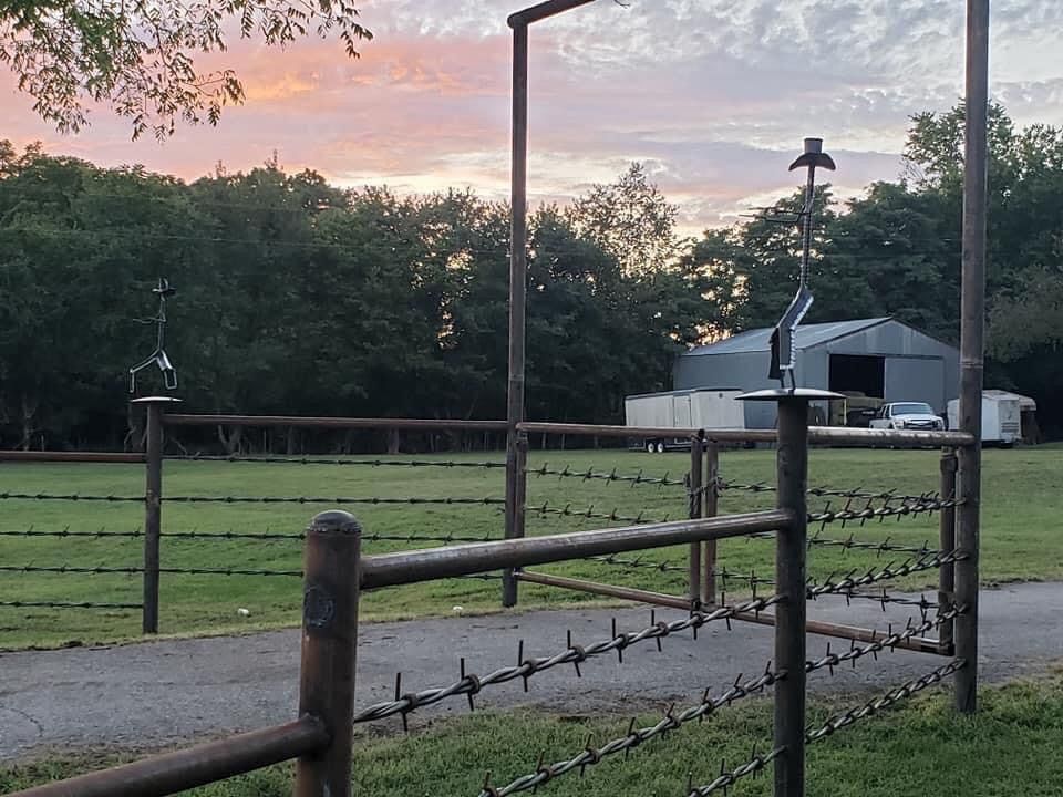 Barbed-wire fence and gate beside a gravel drive at sunset, with a white barn and trees in the background