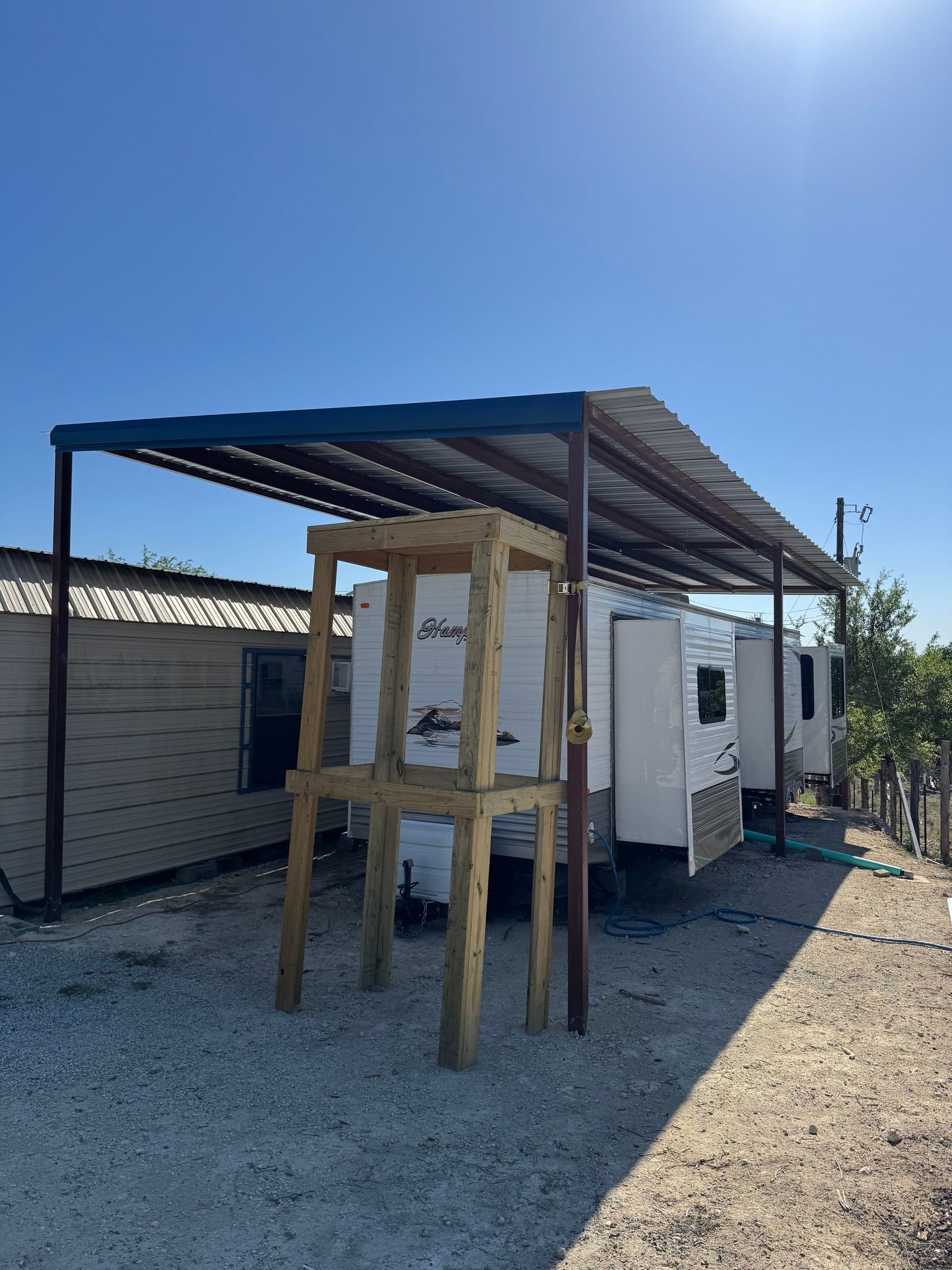 Outdoor workbench under a corrugated metal roof beside a small white shed in a gravel yard