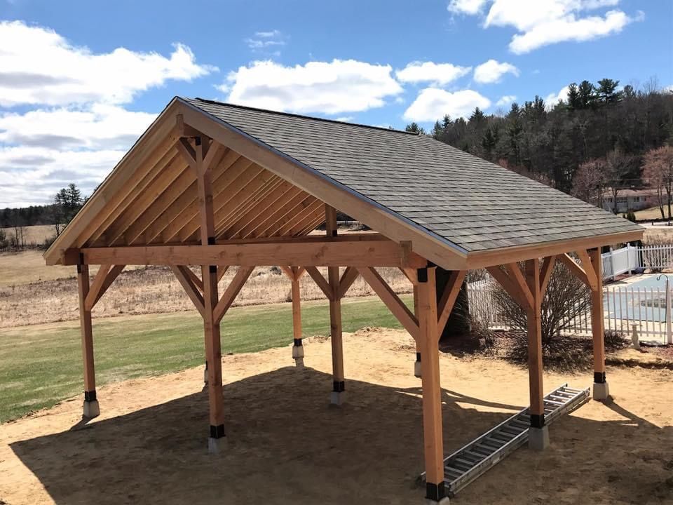 Wooden pavilion with a gray shingled roof on a dirt lot under a partly cloudy sky.