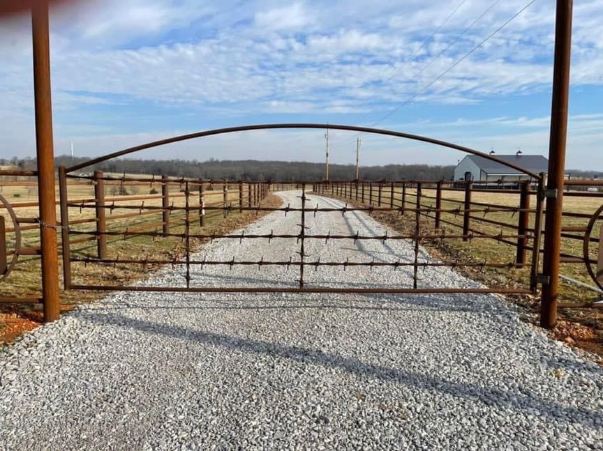 Gravel driveway leads to a metal gate and fence under a blue sky in a rural field