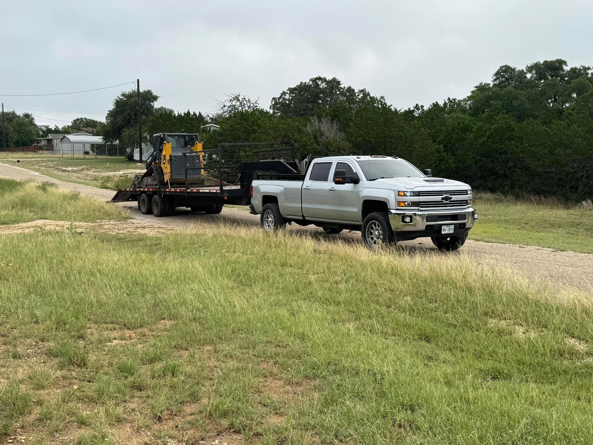 White pickup towing a backhoe on a flatbed trailer along a dirt road beside green trees