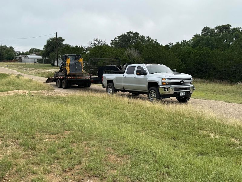 Silver pickup truck towing a skid steer on a flatbed trailer along a gravel path by trees