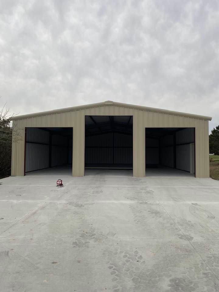 Large beige metal garage with three open bays on a concrete lot under a cloudy sky.