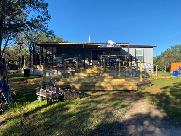 Wooden deck with stairs on a house, with screened porch, trailer, and trees in a sunny yard.