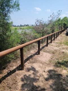 Rusty metal railing along a dirt path beside a sunlit green field and trees