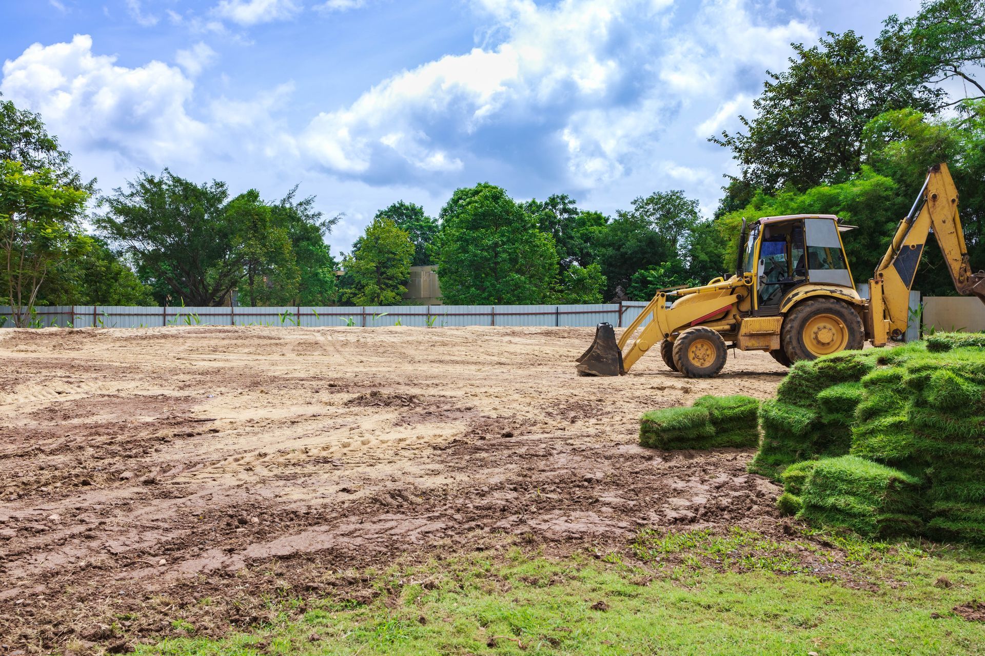 Yellow backhoe on muddy field beside piled sod under a blue sky