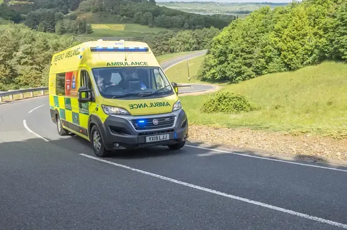 A yellow emergency ambulance with blue flashing lights drives along a curving road in a green, hilly landscape.