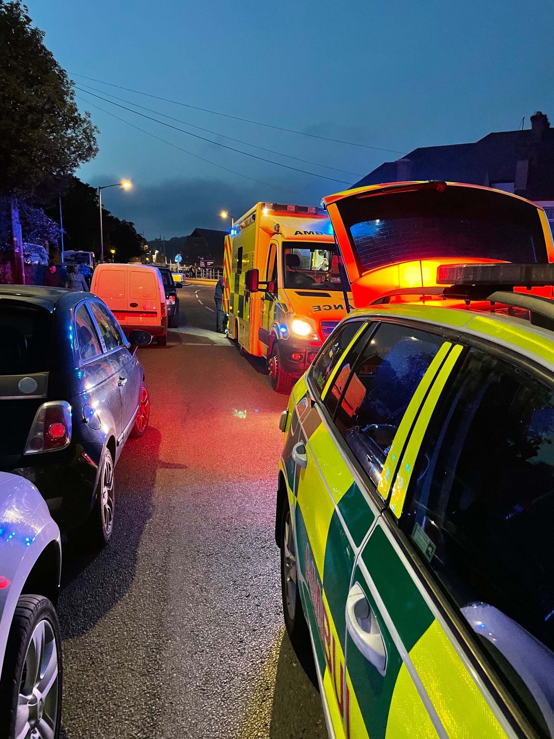 Emergency vehicles with flashing lights parked on a road at twilight with other vehicles nearby.
