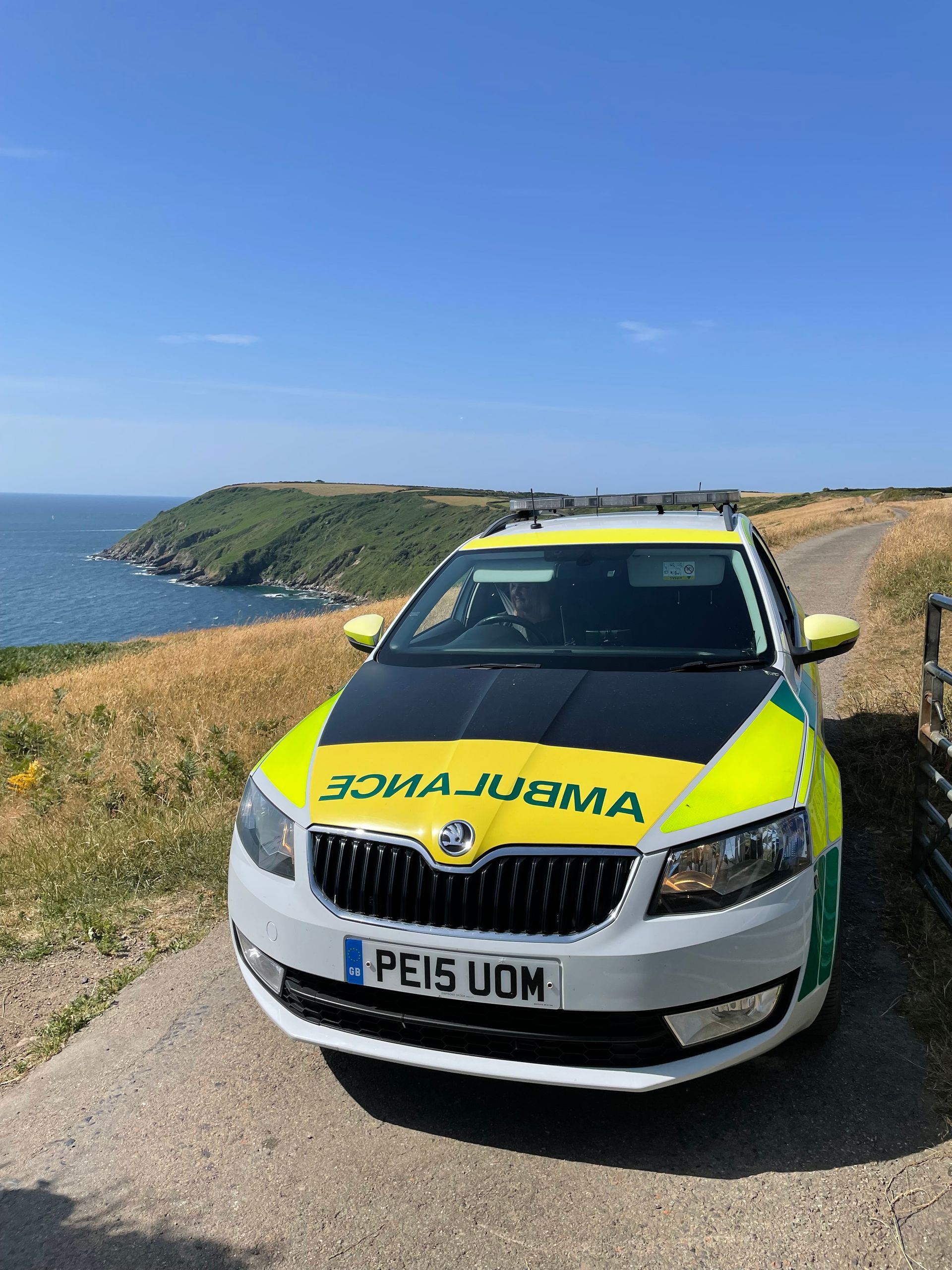 A yellow and green Skoda ambulance parked on a coastal path overlooking the sea under a clear blue sky.