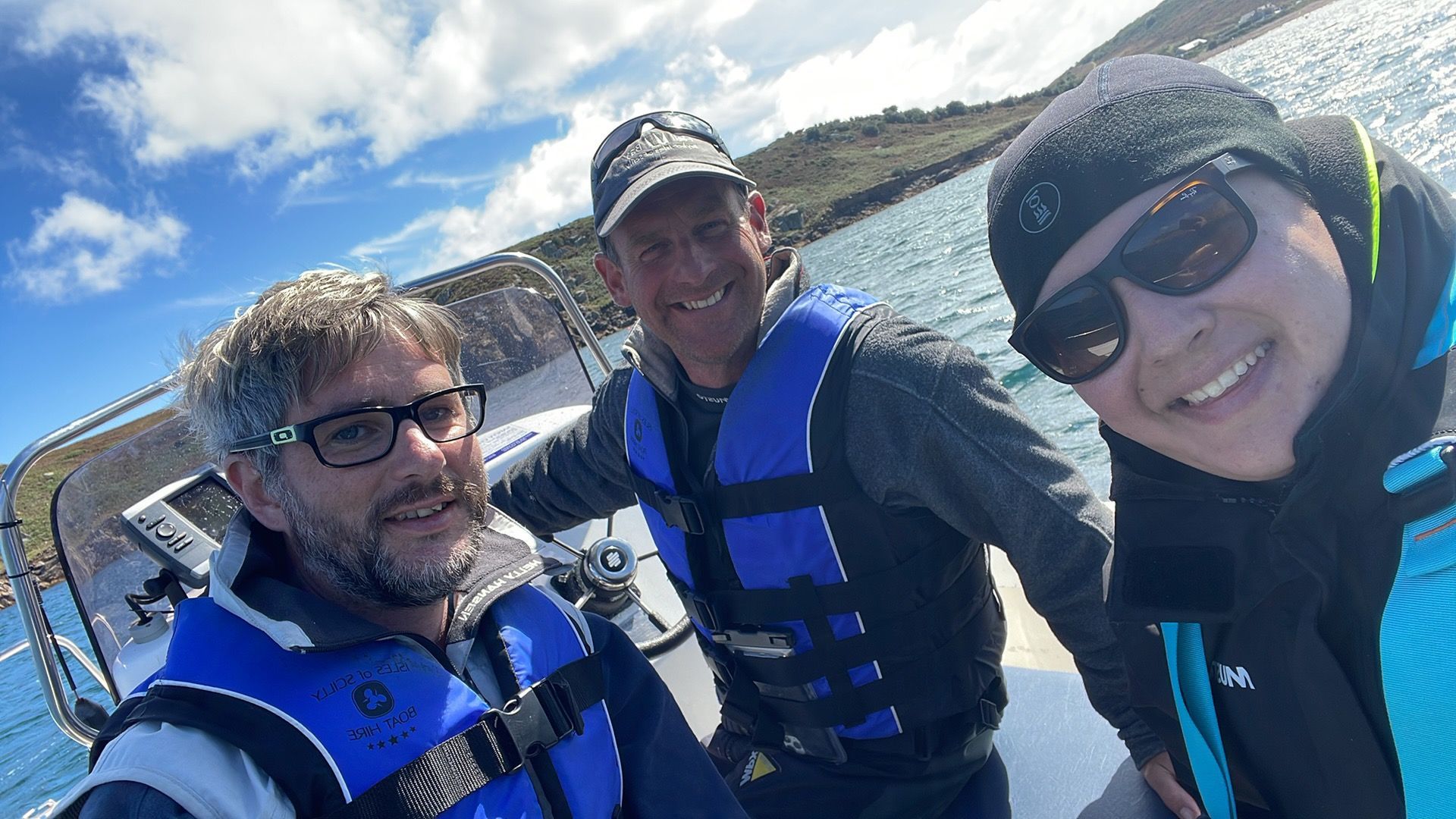 Three people wearing life jackets smile together on a boat under a bright, partly cloudy sky.