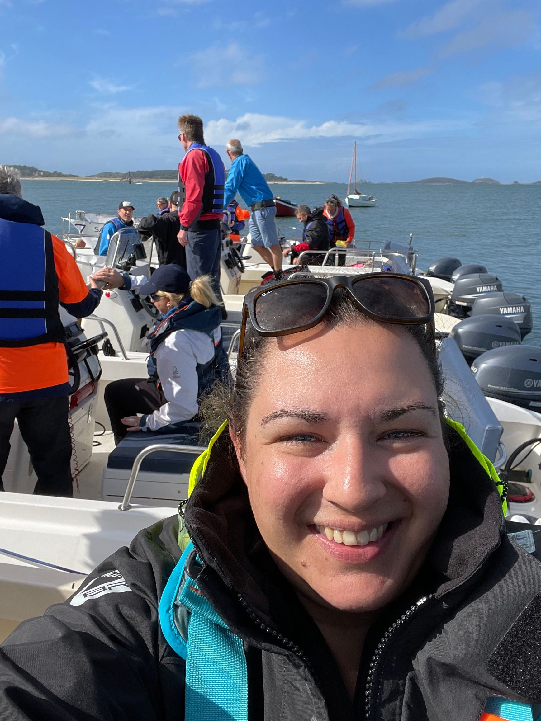 A smiling person takes a selfie on a boat filled with passengers wearing life jackets on a sunny day at sea.